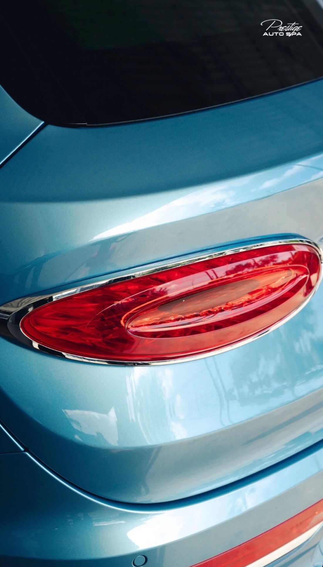 Oval red tail light on a pale blue Bentley with glossy paint reflecting the sky